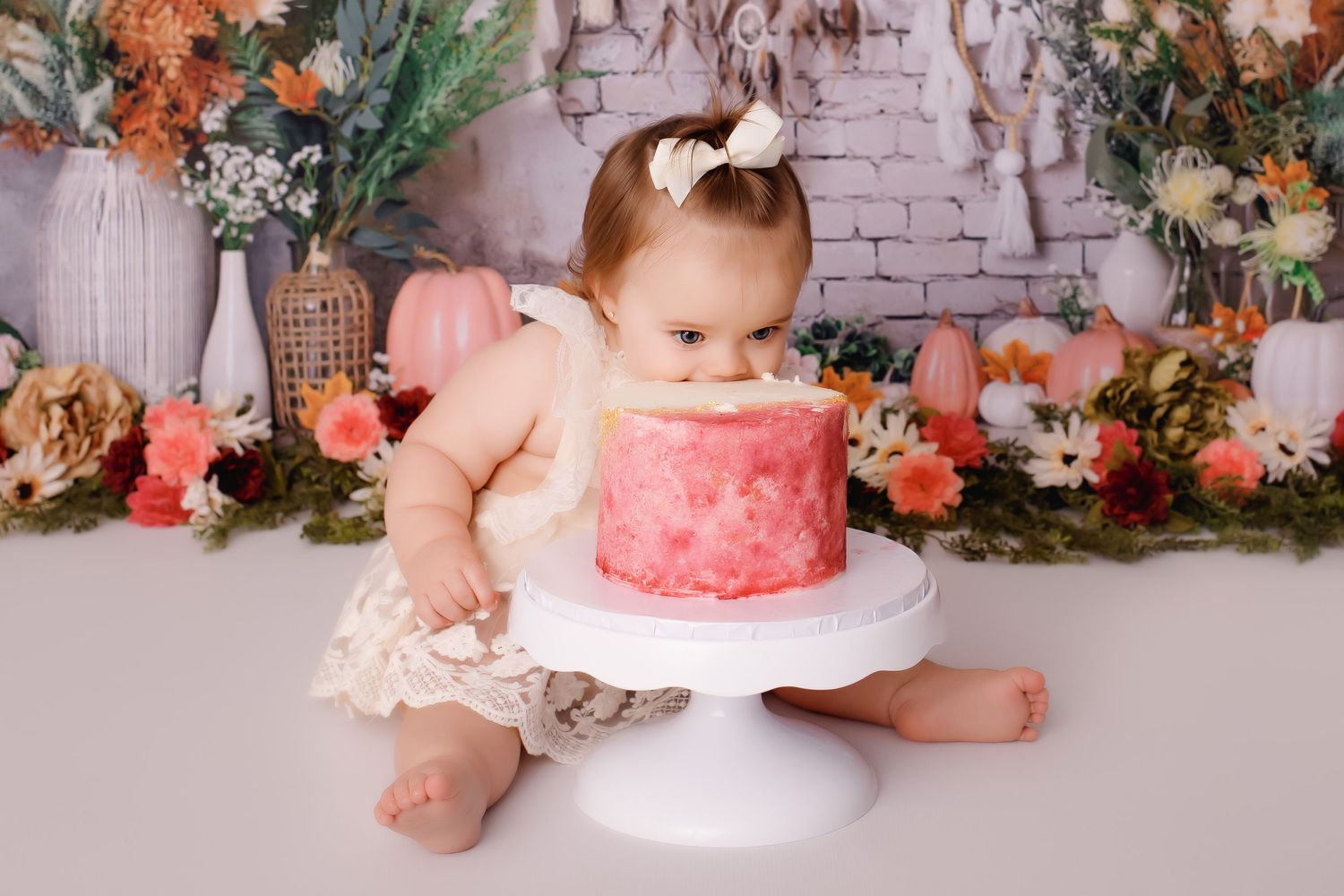 A baby in a white dress reaching for a pink cake on a pedestal against a floral backdrop.