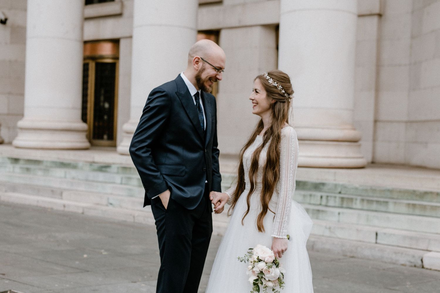 A wedding couple shares a joyful moment between classical columns of a grand building.