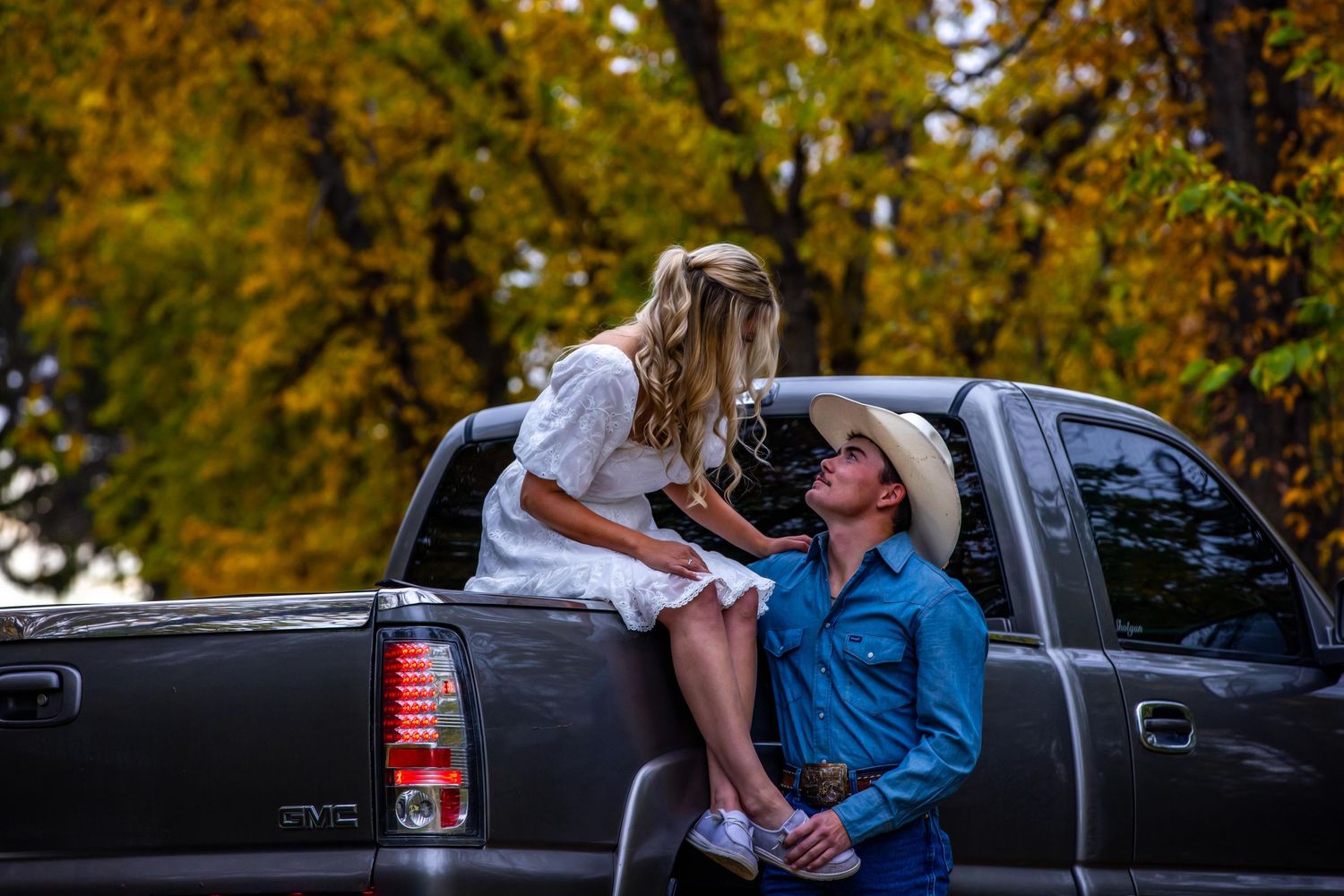 A romantic couple sitting together on a pickup truck tailgate surrounded by fall foliage.