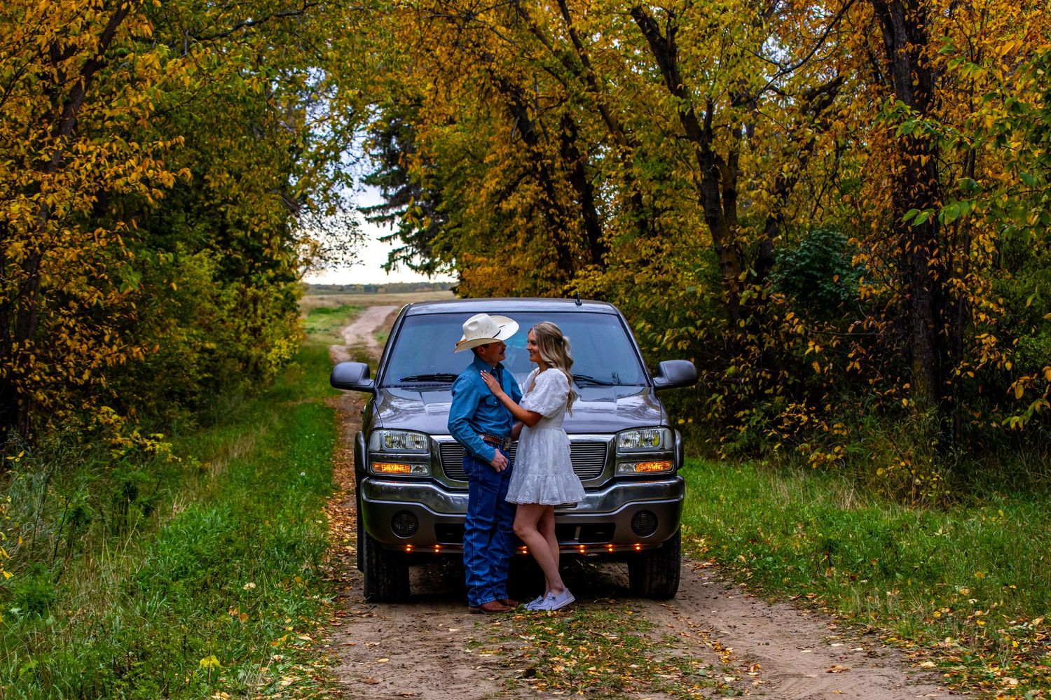 A couple embracing in front of a black pickup truck on a tree-lined dirt road in autumn.