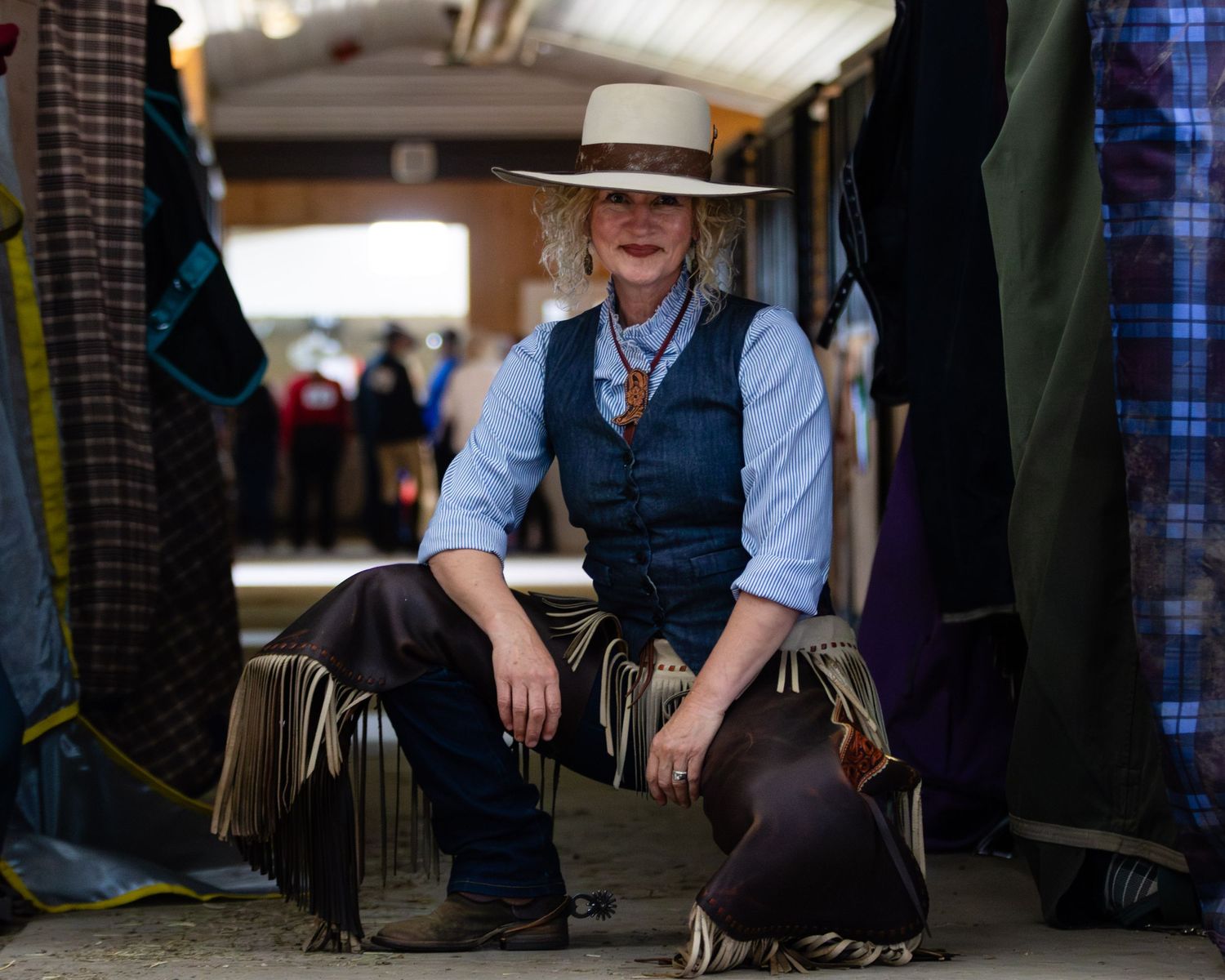 Western-styled figure in vest and hat resting in indoor stable setting.