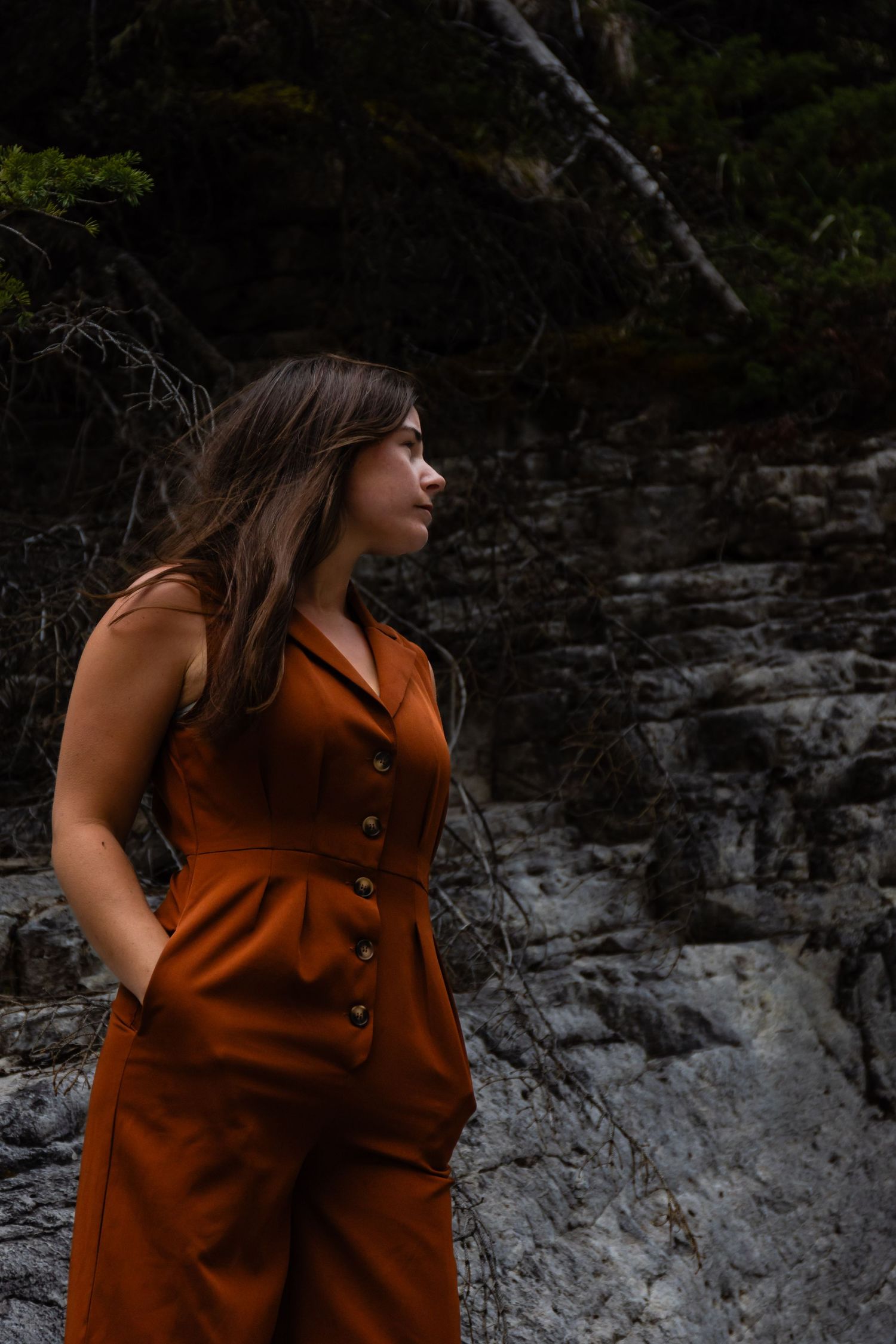A person in a rust-colored button-up jumpsuit poses against dramatic rocky cliffs.