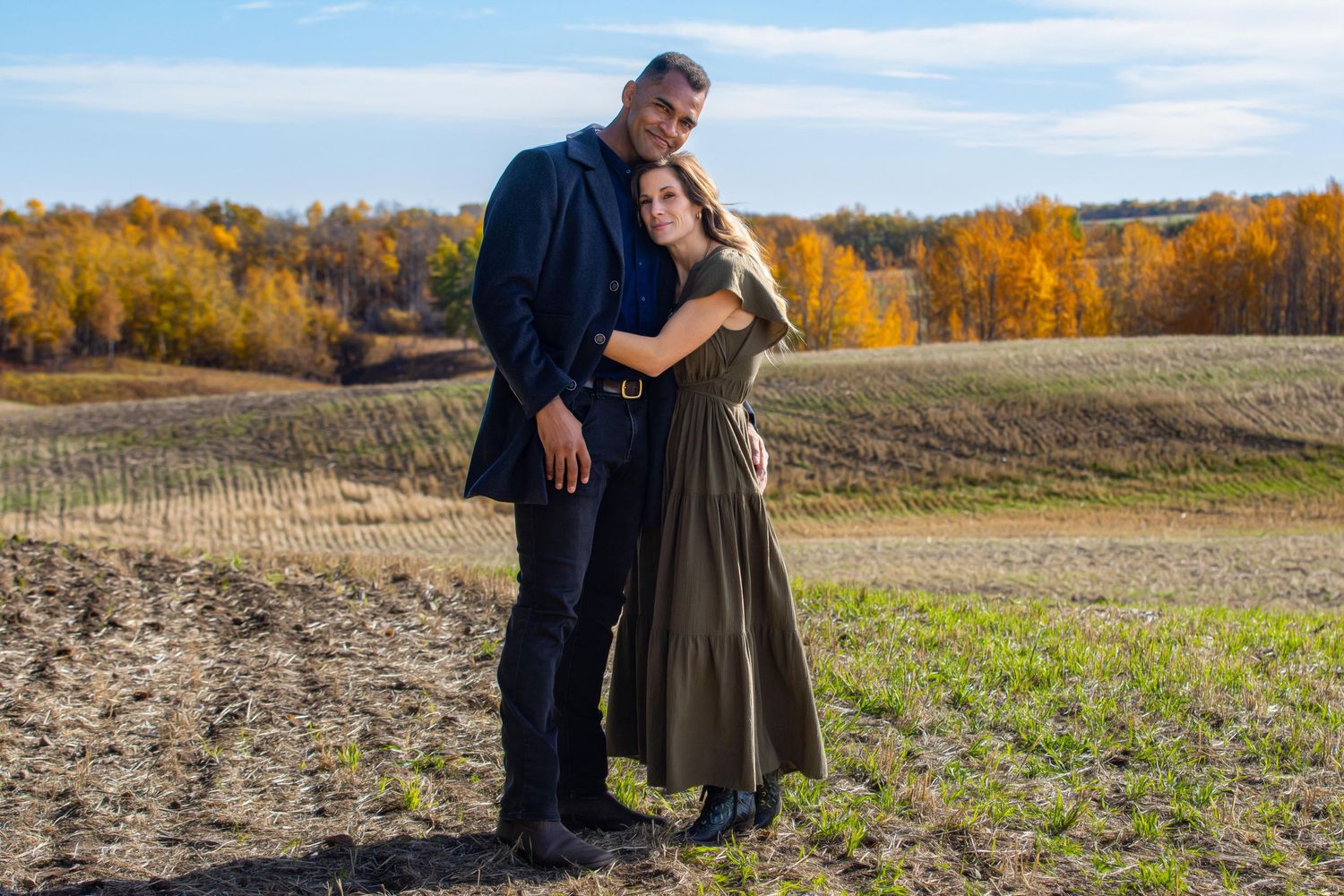 A couple embraces on a dirt path surrounded by autumn trees and fields on a sunny day.