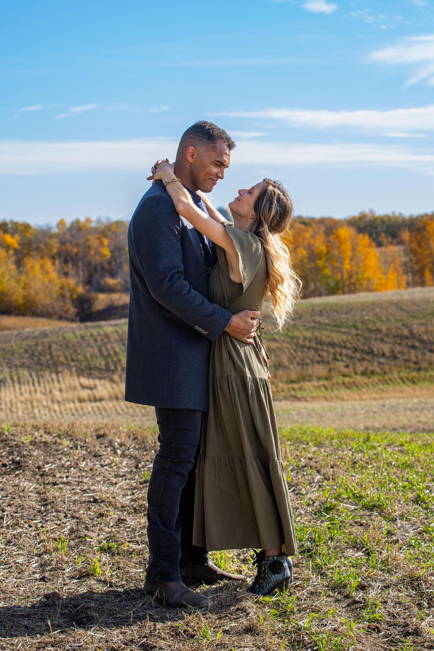 A couple embraces in an autumn field with golden foliage and rolling hills in the background on a sunny day.