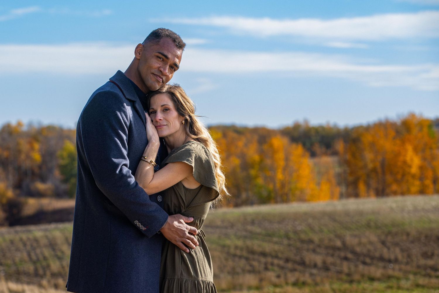 A couple embraces while standing in front of an autumn field with golden trees and blue skies in the background.