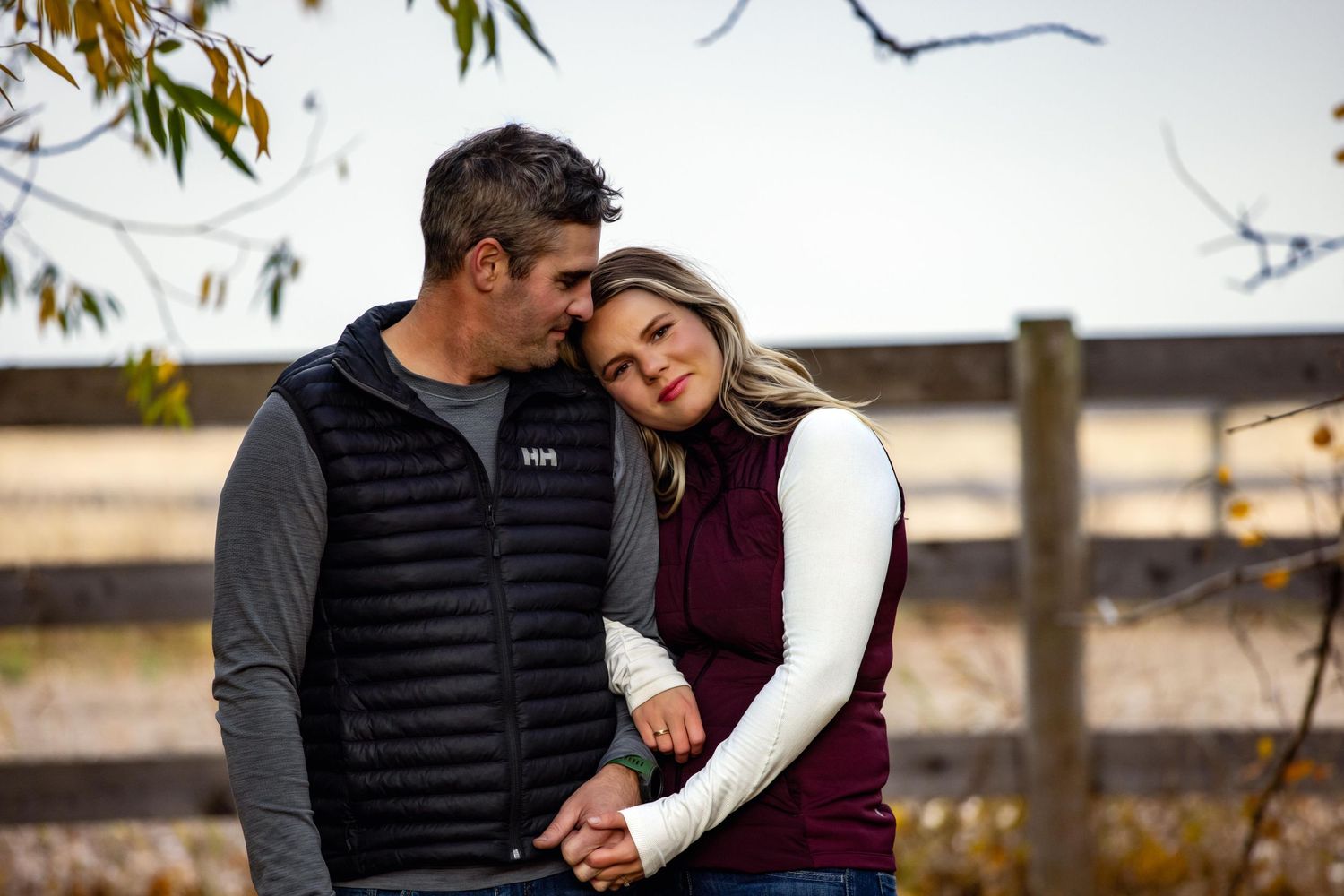 A couple shares a tender moment outdoors while standing near a wooden fence during autumn.