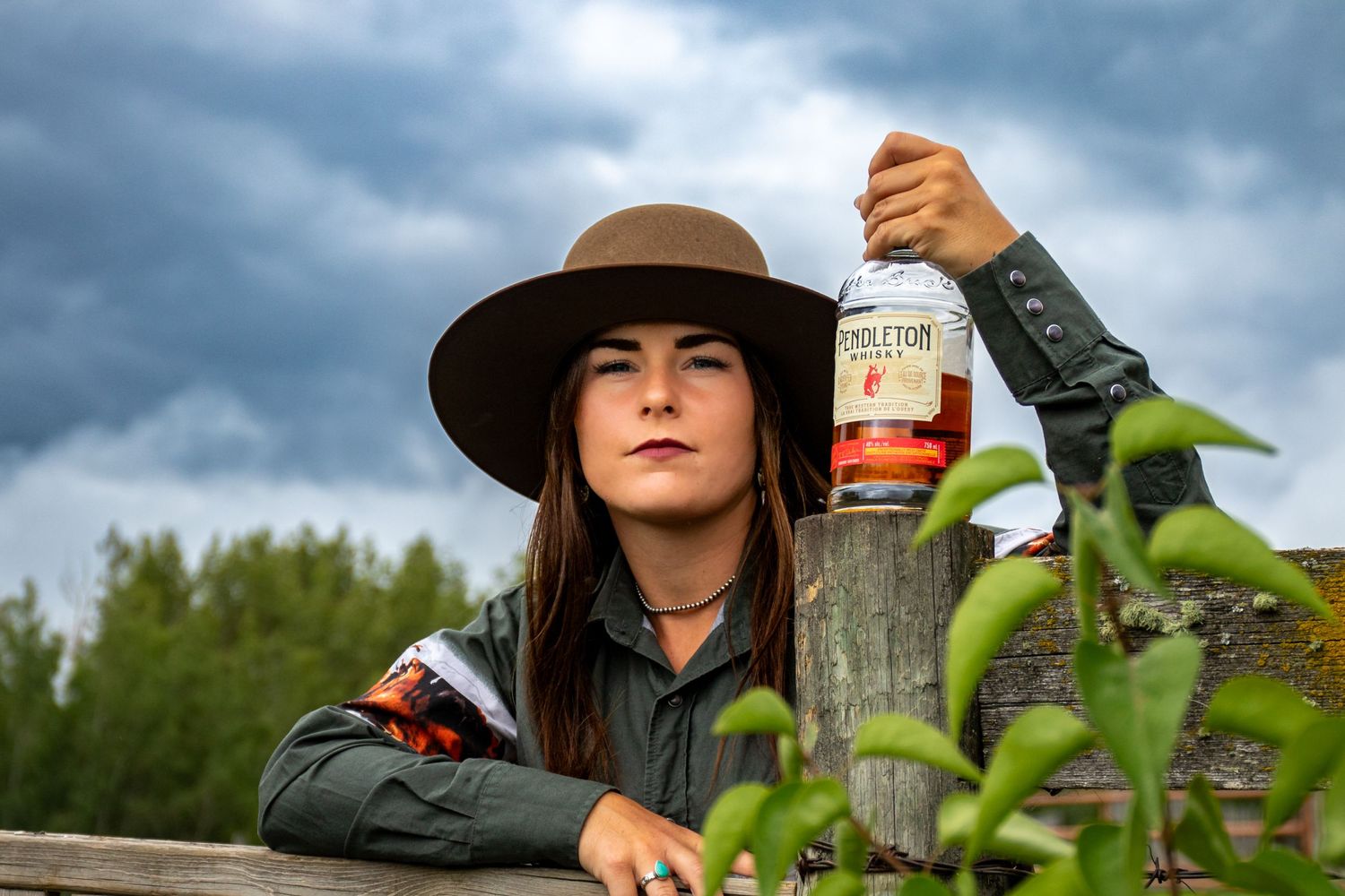 Someone in a wide brim hat poses with a bottle against a stormy sky background.