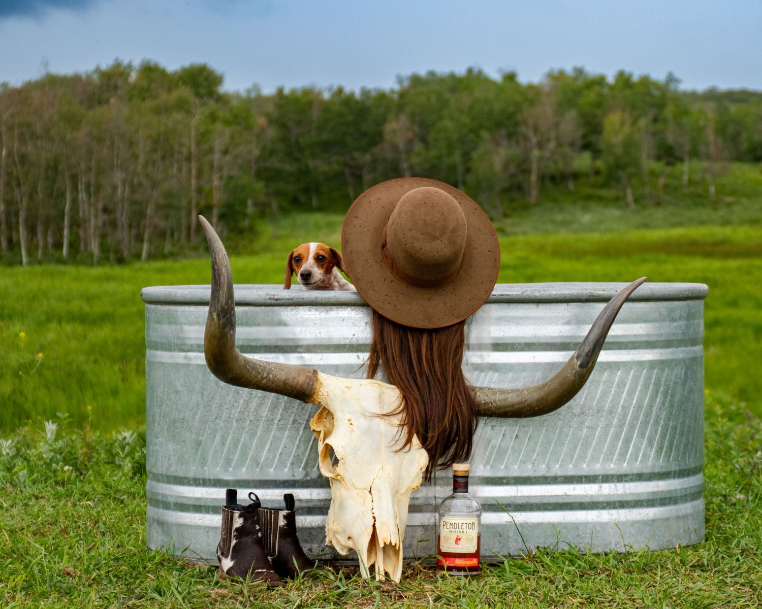A rustic scene with a stock tank, cowboy hat, animal skull and small dog in a meadow.