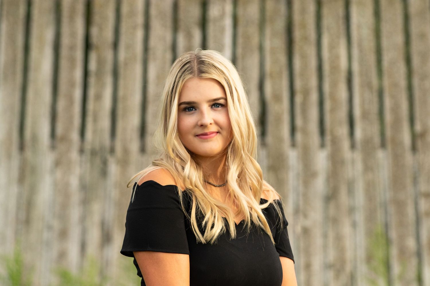A casual portrait in front of wooden fence wearing black off-shoulder top.