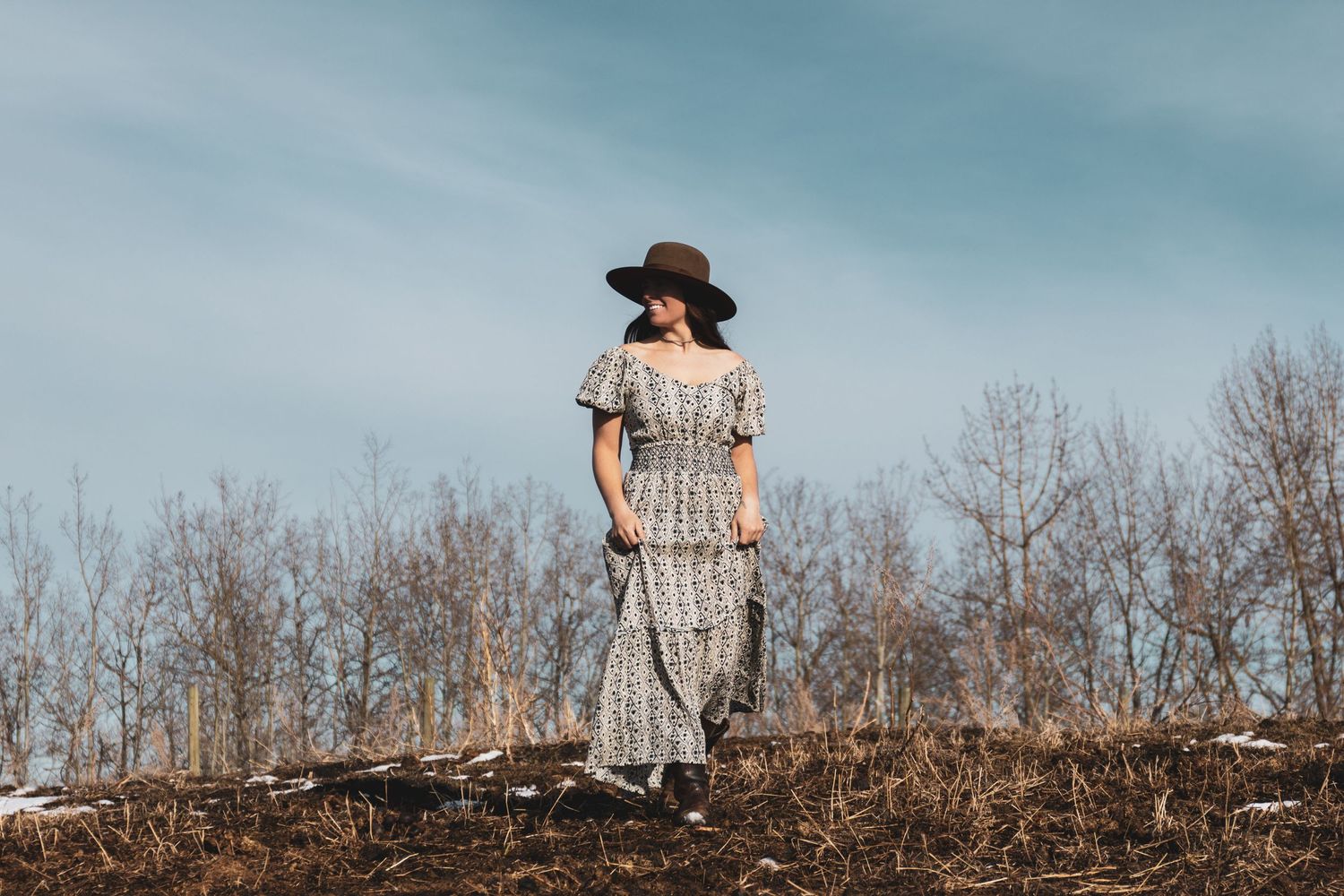 A figure in a vintage dress and hat stands alone on a hilltop against a blue sky and bare trees.
