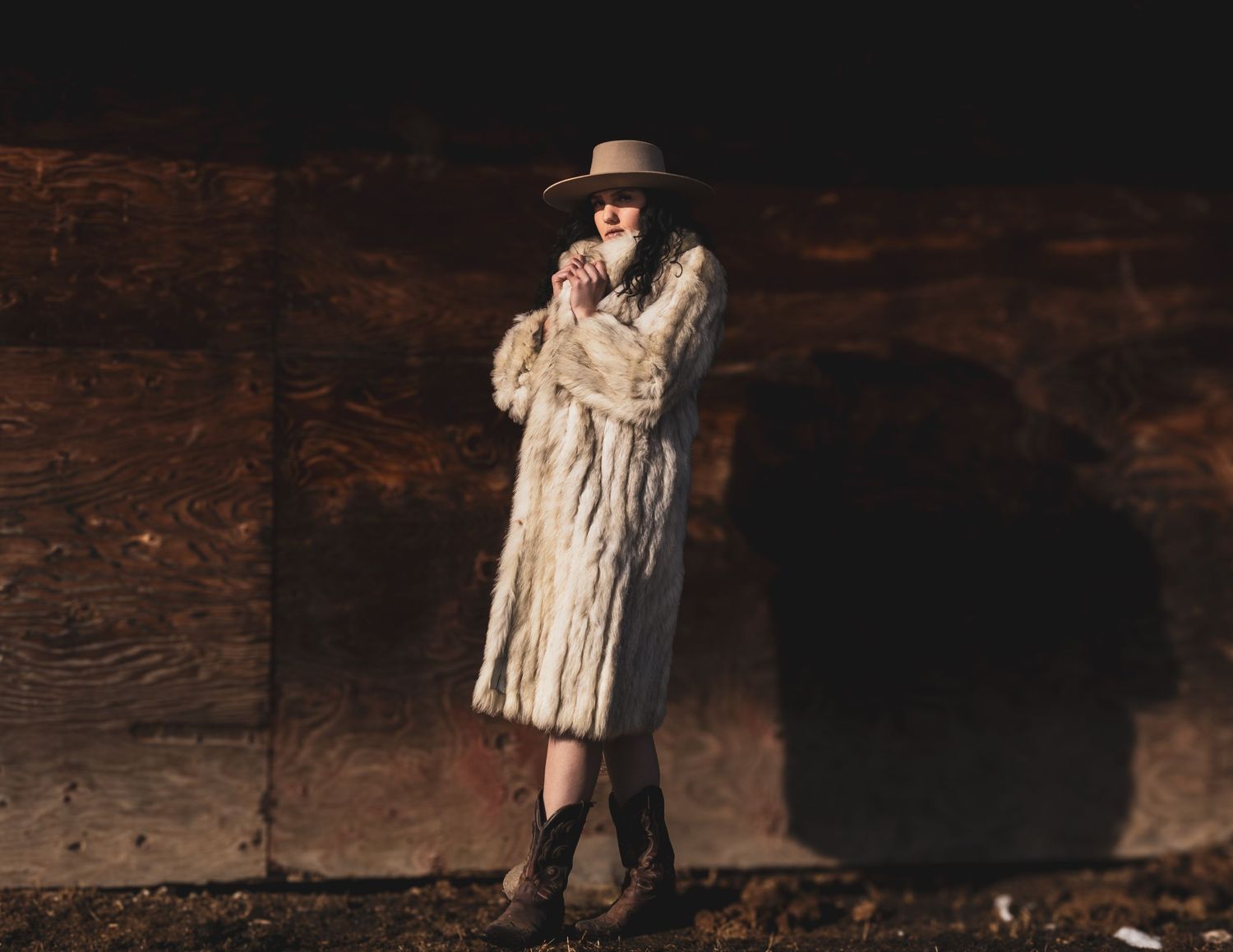 Figure in light-colored coat and hat posed against dark wooden wall.