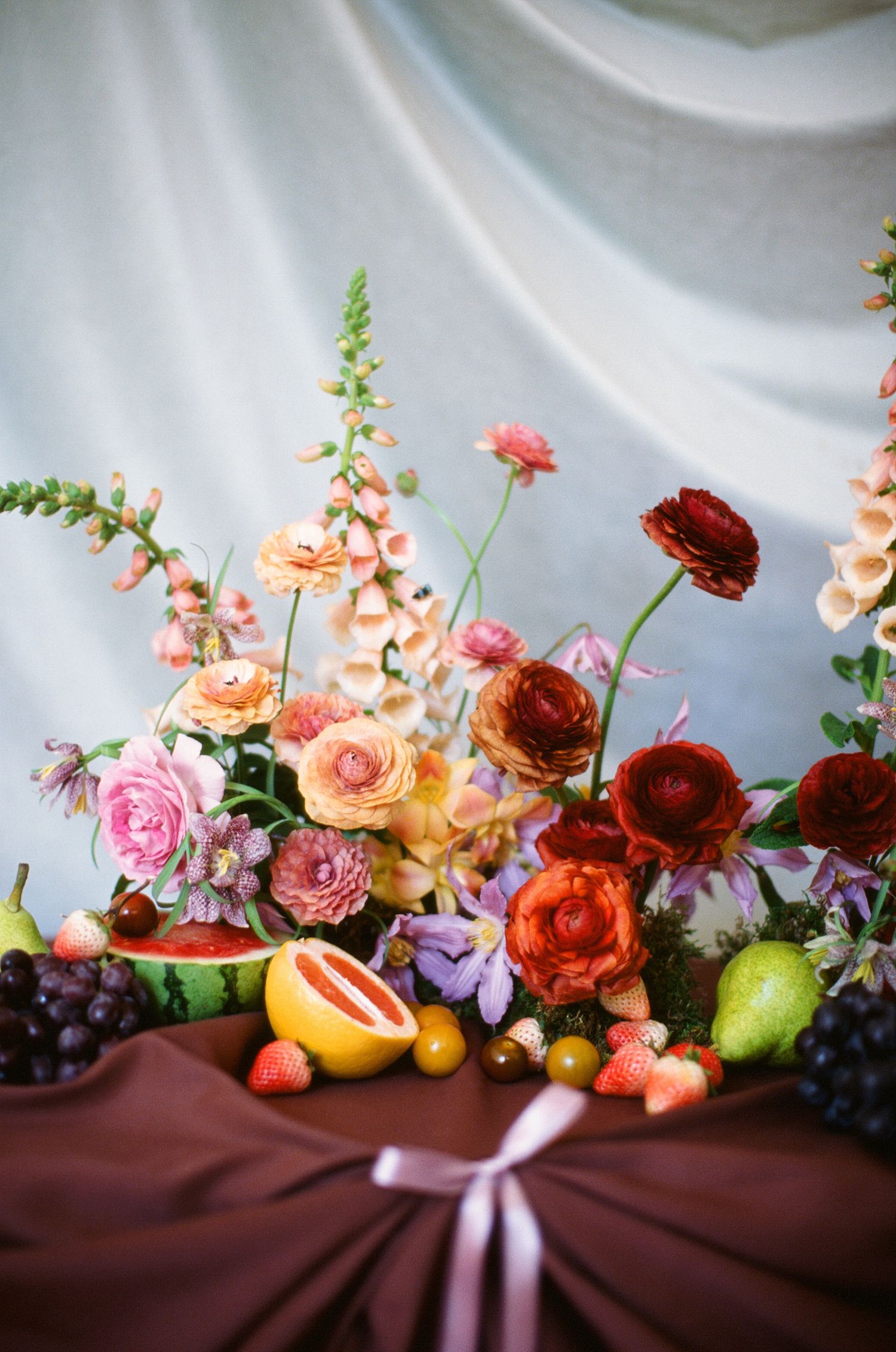 Vibrant floral arrangement with ranunculus, roses, and citrus fruits against white backdrop.