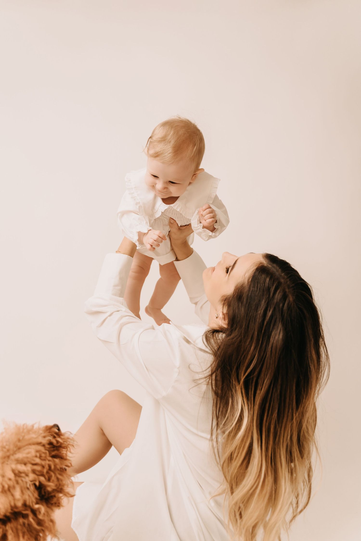 Mother in white dress lifts laughing baby against neutral background.