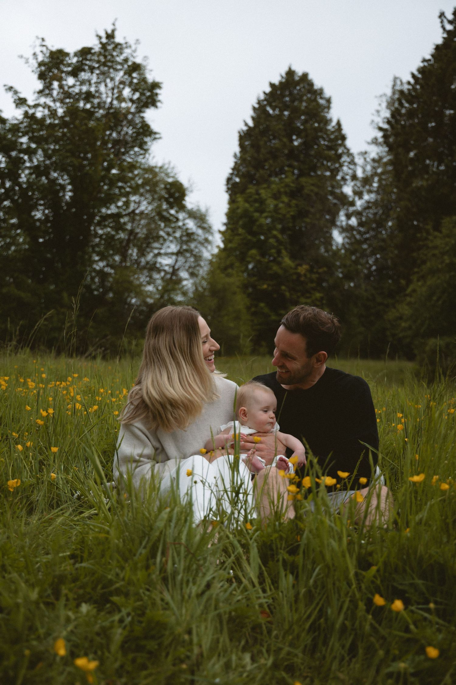 Couple looking at each other while holding their six months old baby sitting in a yellow flower field on a rainy day in Vancouver, BC.