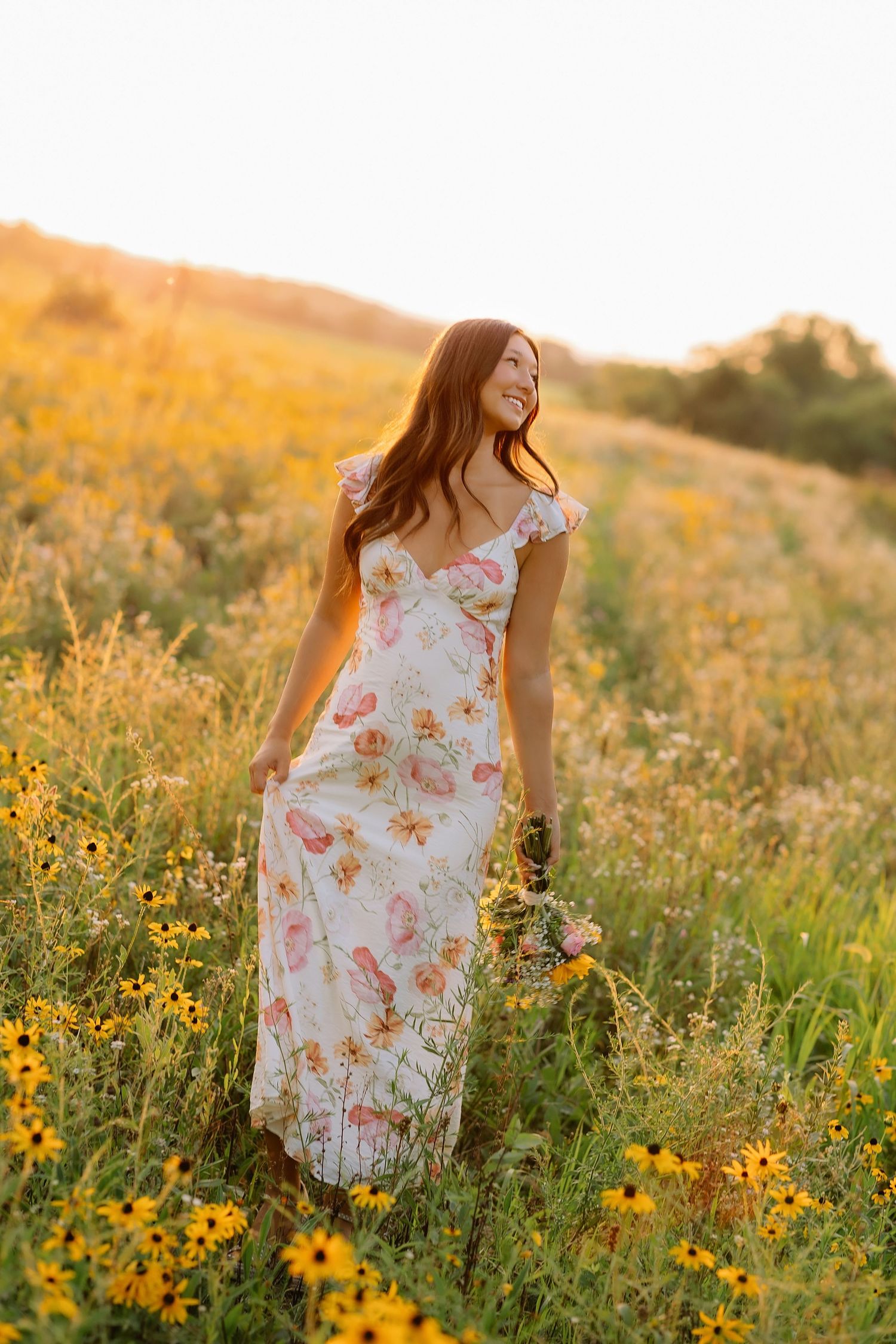 Young woman in bohemian outfit with cowboy boots sits by a rustic fence at golden sunset on a farm.