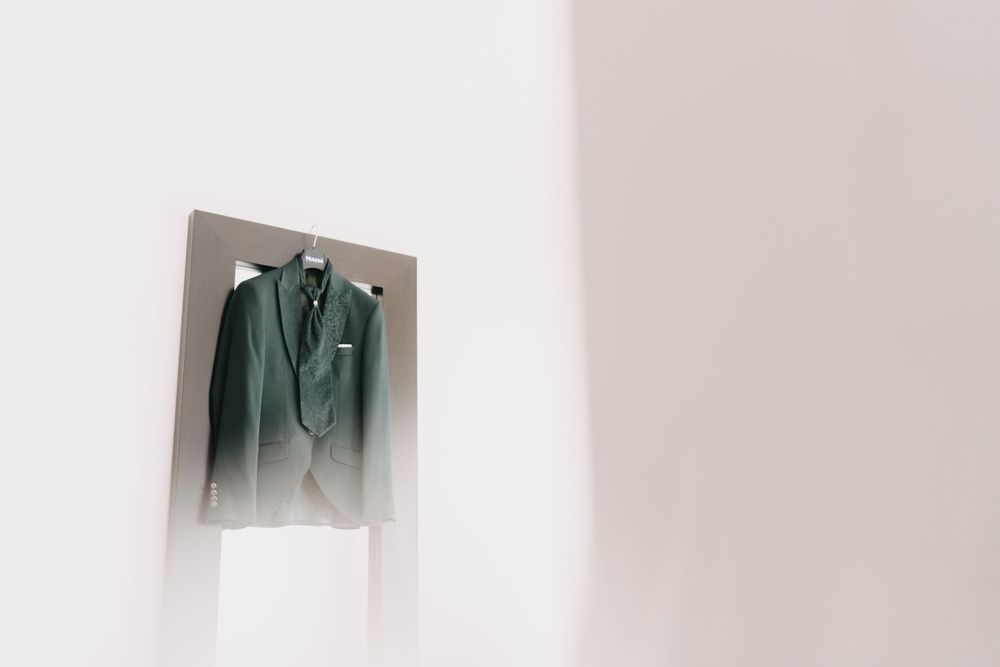 Green formal suit jacket displayed on hanger against minimalist white wall.