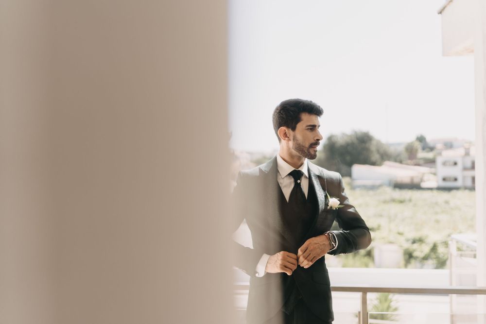 Elegant groom in formal attire looks pensively through window during wedding preparation.