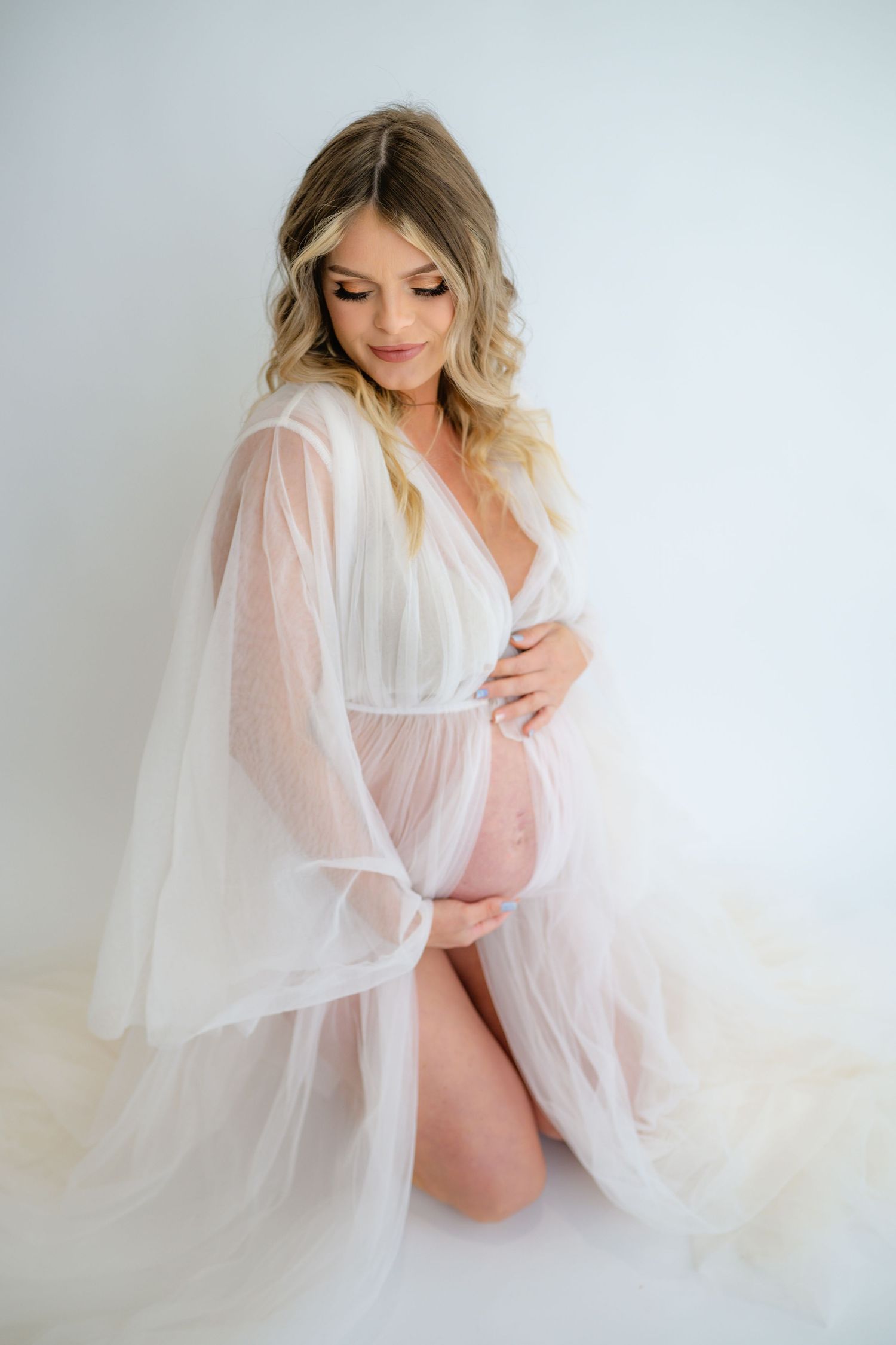 Ethereal maternity photo in flowing white sheer robe against light background showcasing pregnancy glow.