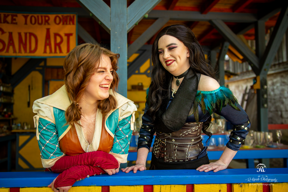 Two cosplayers in medieval-inspired outfits share a moment of laughter at a sand art station.