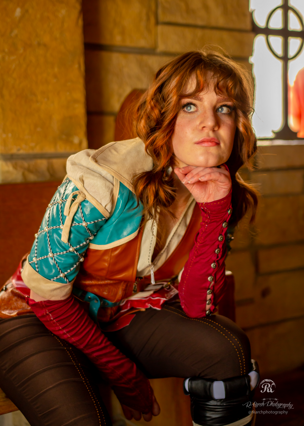 A cosplayer in a Renaissance-inspired outfit with turquoise and brown leather elements poses thoughtfully by a window.