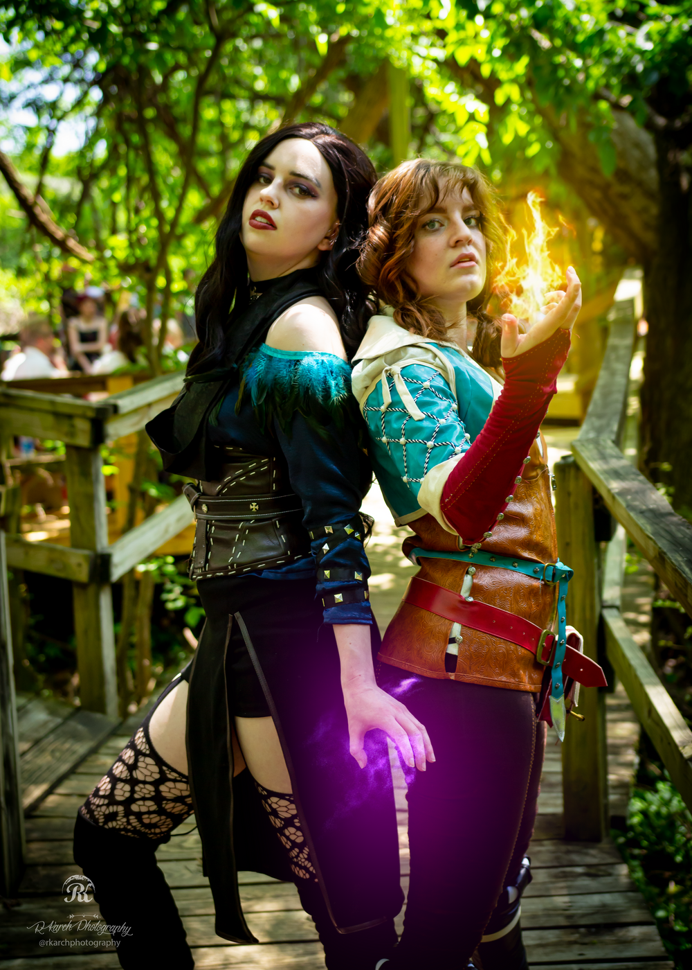 Two cosplayers in contrasting dark and light fantasy outfits pose back-to-back on a wooden bridge.
