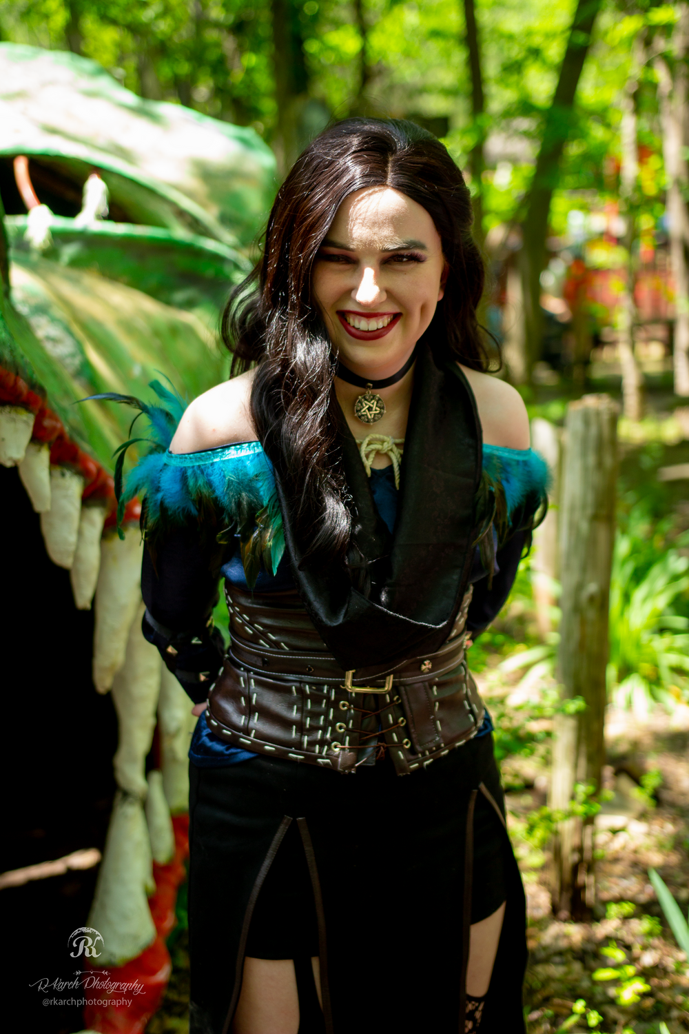 A cosplayer in a steampunk-inspired outfit with teal accents poses joyfully in a wooded setting.