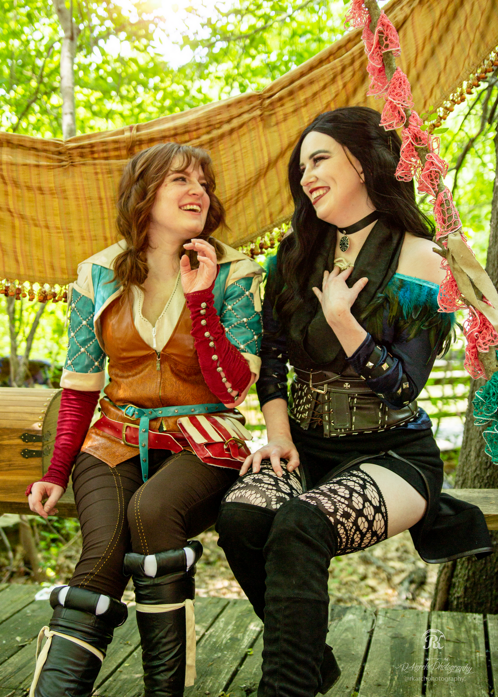 Two cosplayers in fantasy-themed costumes share a laugh while sitting together under a canopy.