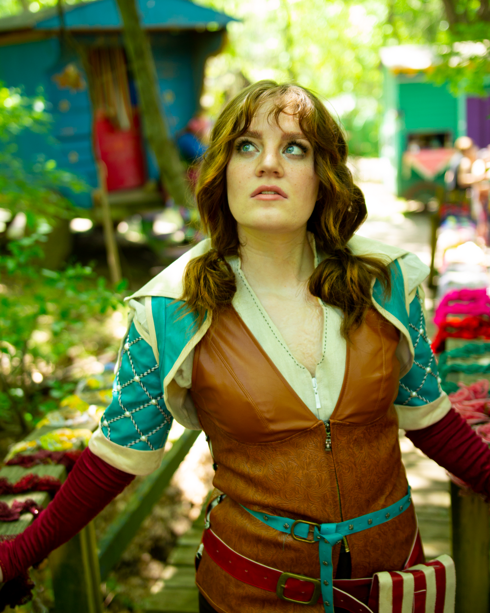 A cosplayer in a brown leather vest and teal sleeves poses outdoors with a curious expression.
