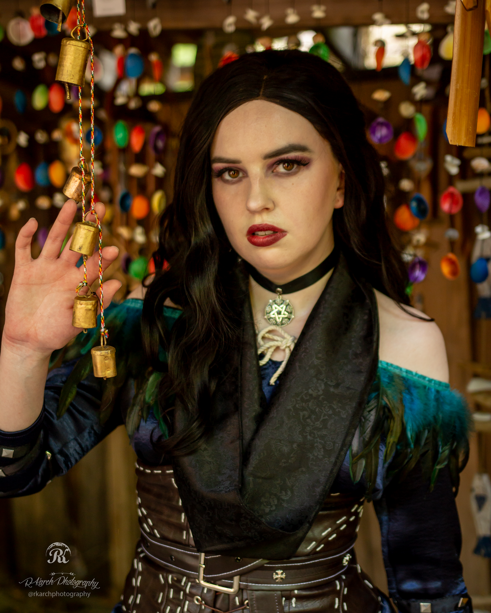 A gothic-styled cosplayer in a corset and off-shoulder dress poses with decorative bells against colorful lights.