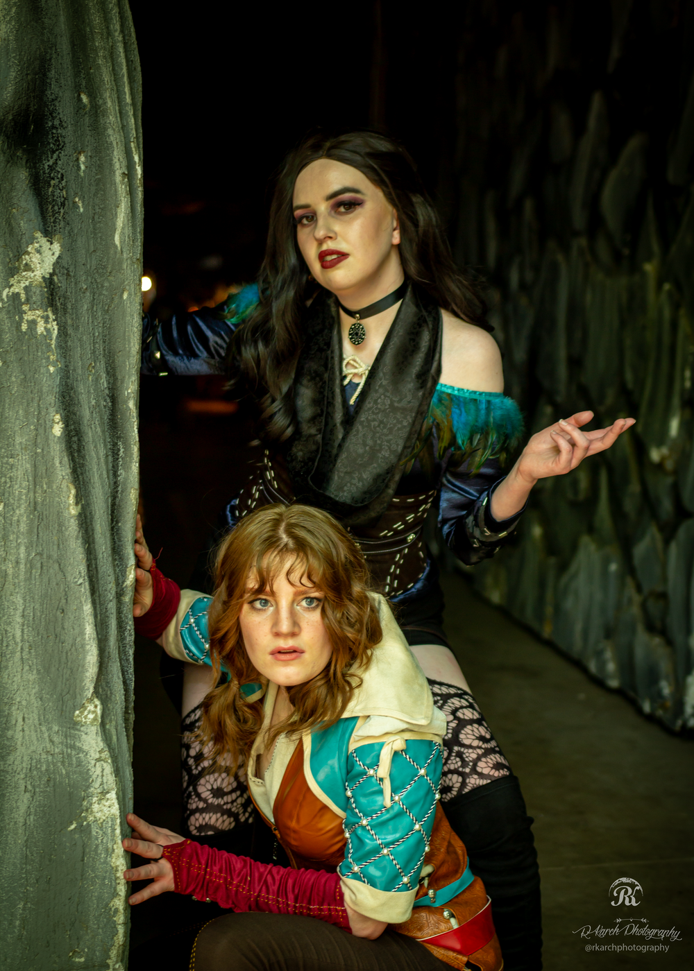 Two cosplayers in fantasy attire pose dramatically in a dark corridor setting.