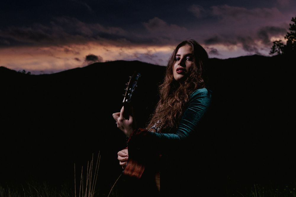 A musician in a teal sweater holds a guitar against a dramatic sunset mountain landscape.