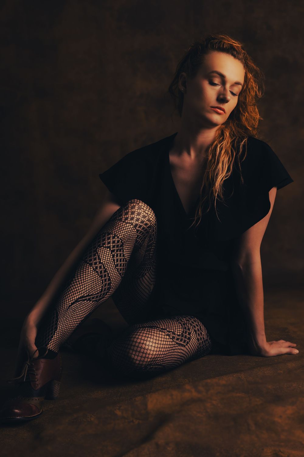 A moody fashion portrait of a figure in black dress with fishnet stockings sitting on dark textured ground in warm lighting.