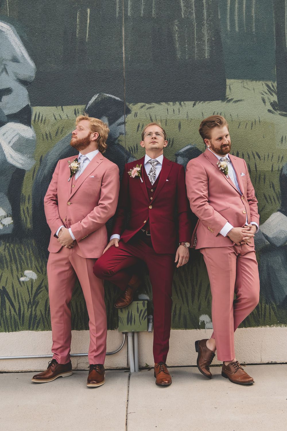 Three groomsmen pose against a mural wearing stylish pink and burgundy suits with brown shoes and floral bow ties.