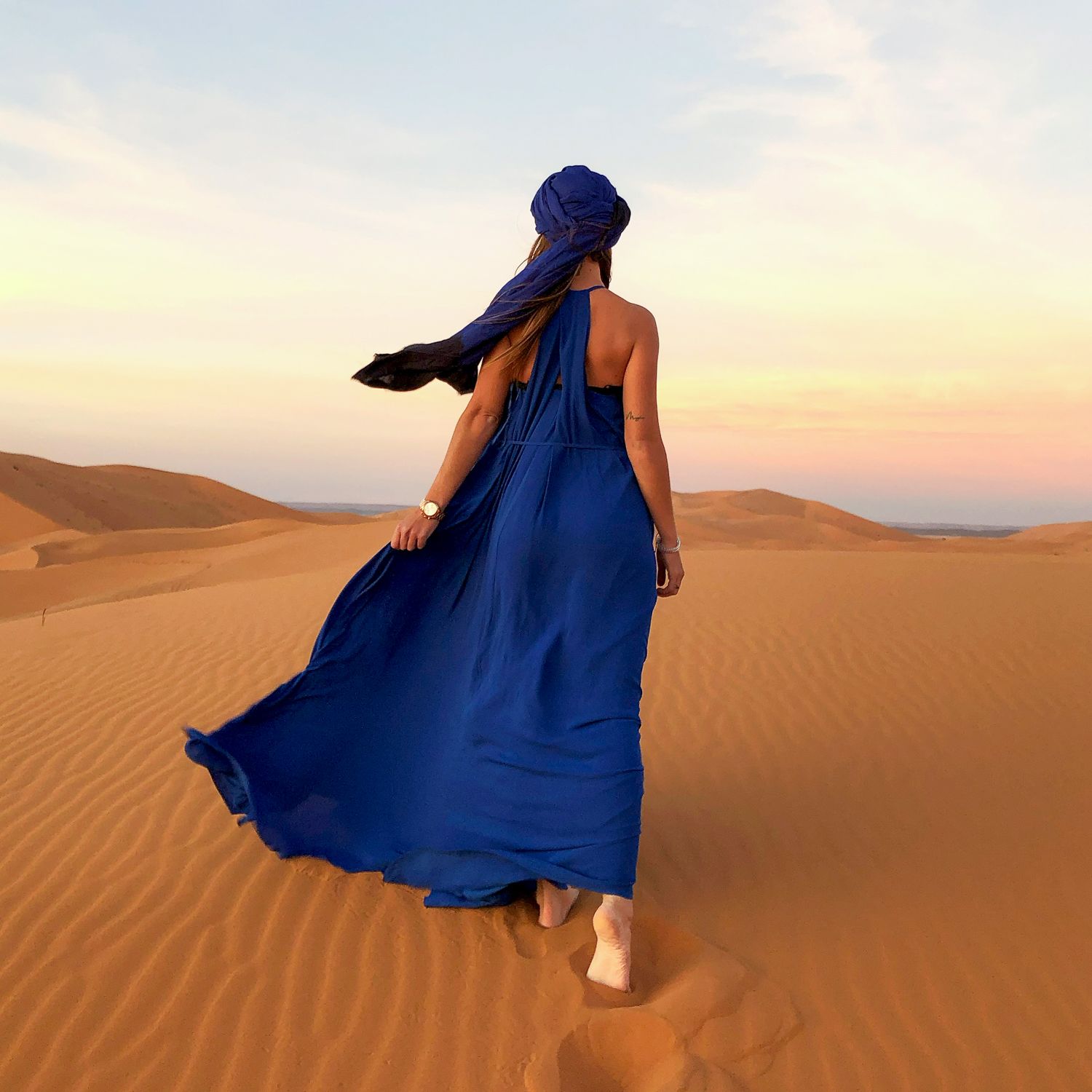 A flowing royal blue dress billows in the desert wind as someone walks across golden sand dunes at sunset.