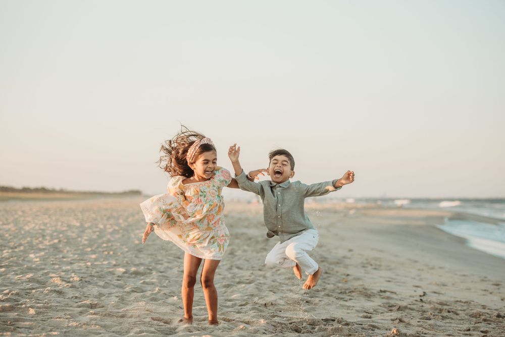 Two children jumping with joy on a sandy beach at sunset with ocean waves in the background.