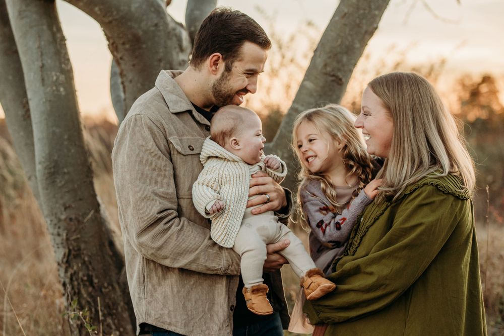 A family shares a tender moment together outdoors at sunset among tree trunks in a natural setting.