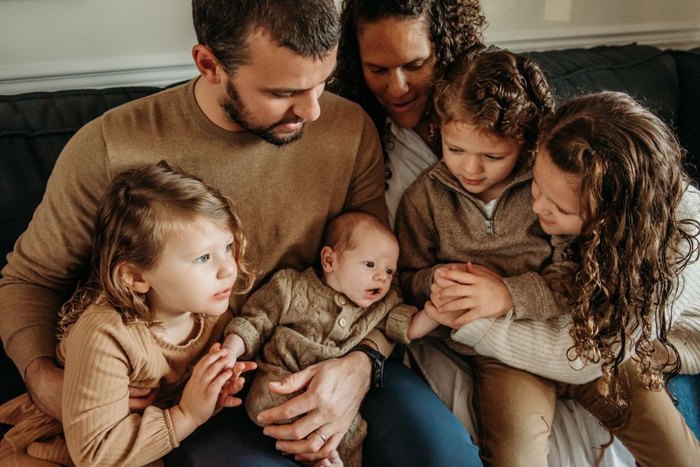 Family of five in matching beige sweaters snuggling together on couch during intimate indoor photo session.