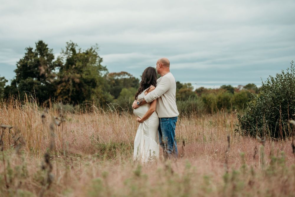 A couple embraces in a rustic field with tall golden grass and trees under an overcast sky.