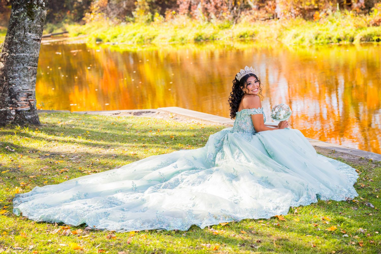 Flowing mint and white gowns spread across grass with golden autumn reflections in water. Quinceañera photos in Pamperin Park.