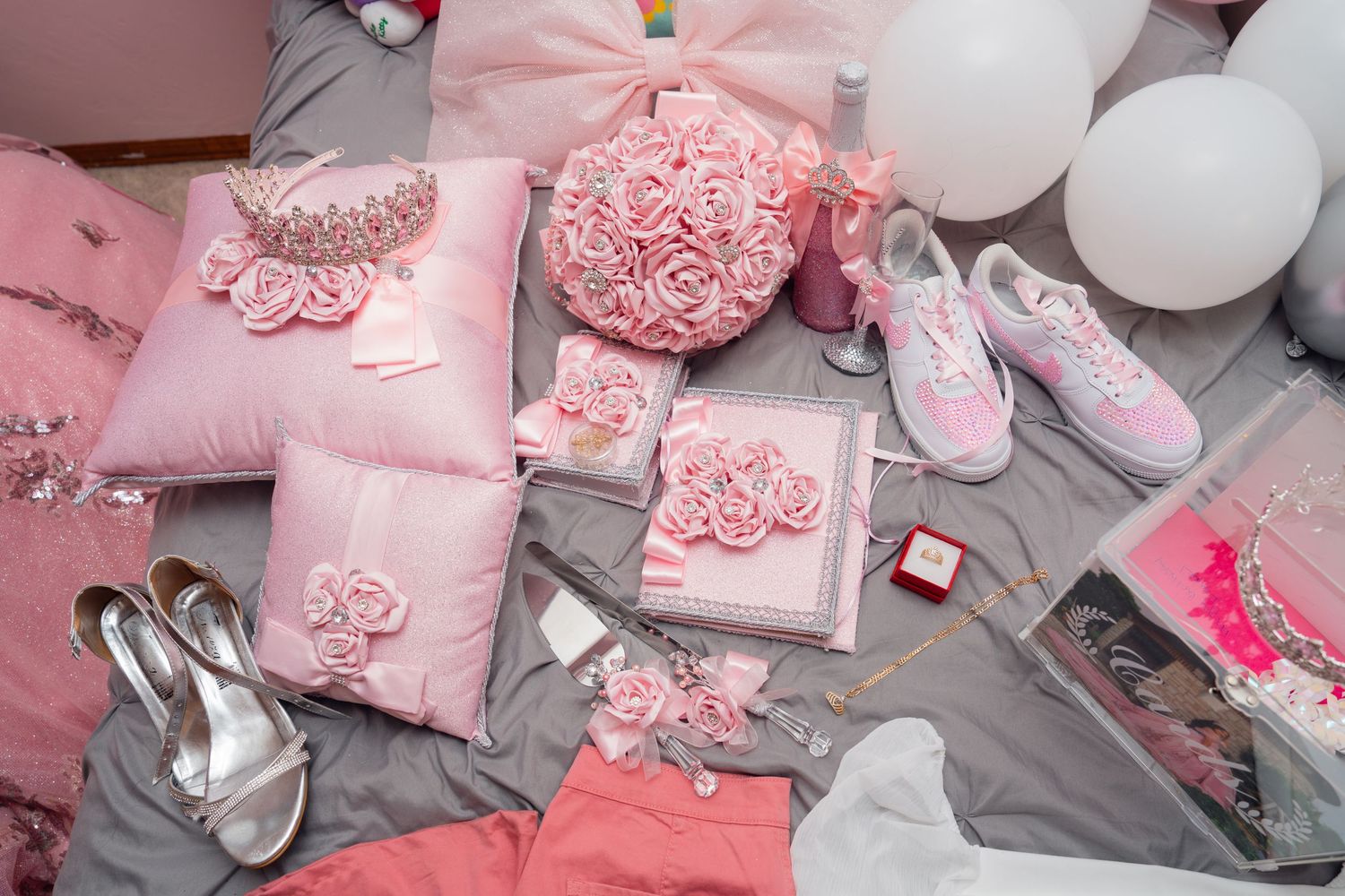 Pink decorative pillows, shoes, and accessories arranged on gray fabric in a flat lay style. Quinceañera