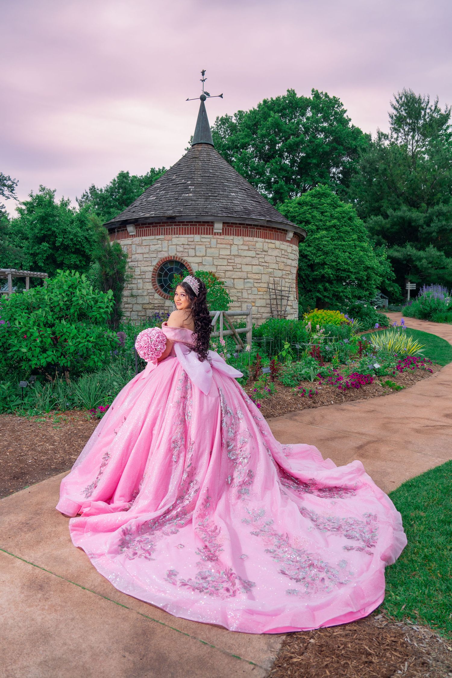 A stunning pink ball gown with intricate embellishments photographed in a garden setting with stone structures. Quinceañera