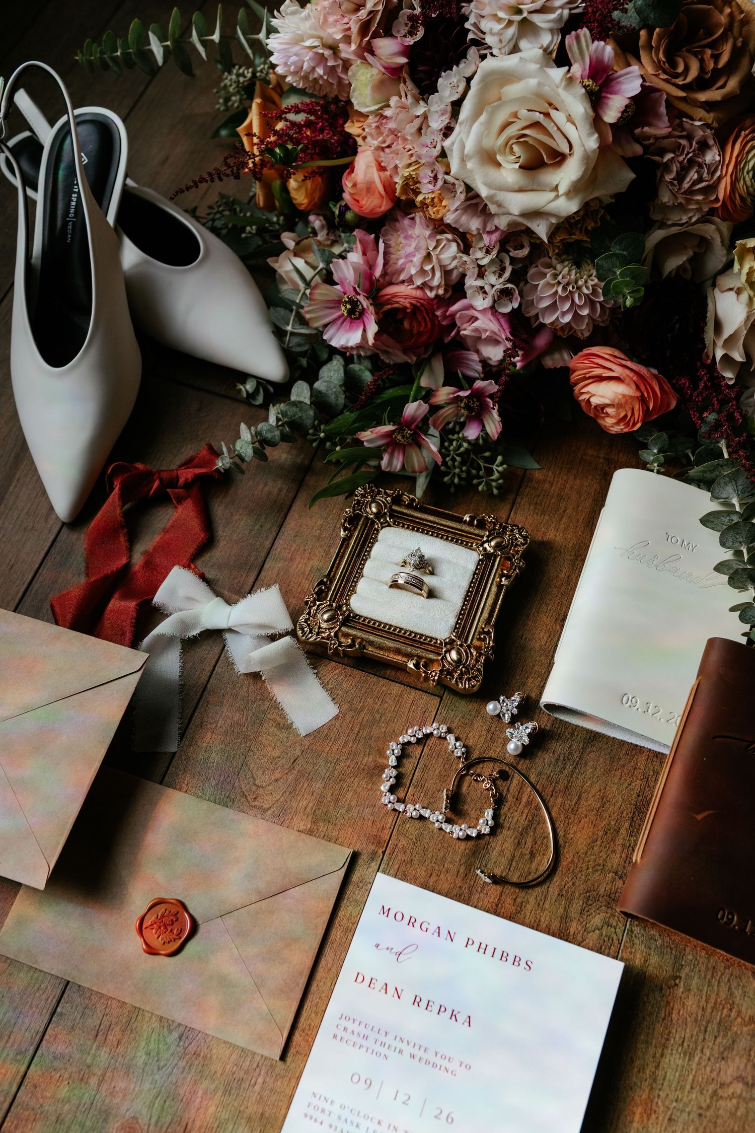 Wedding details including bridal shoes, vow book, jewelry, and perfume arranged on dark wooden surface.