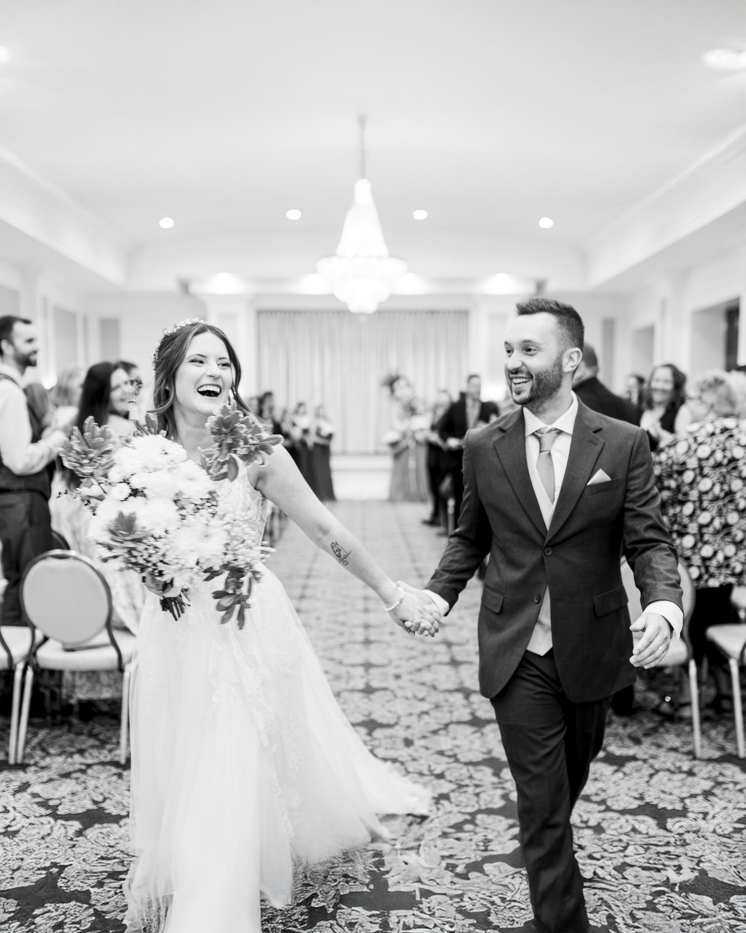 Newlyweds joyfully walk down the Lord Nelson aisle together in an elegant Halifax ballroom with chandelier lighting.