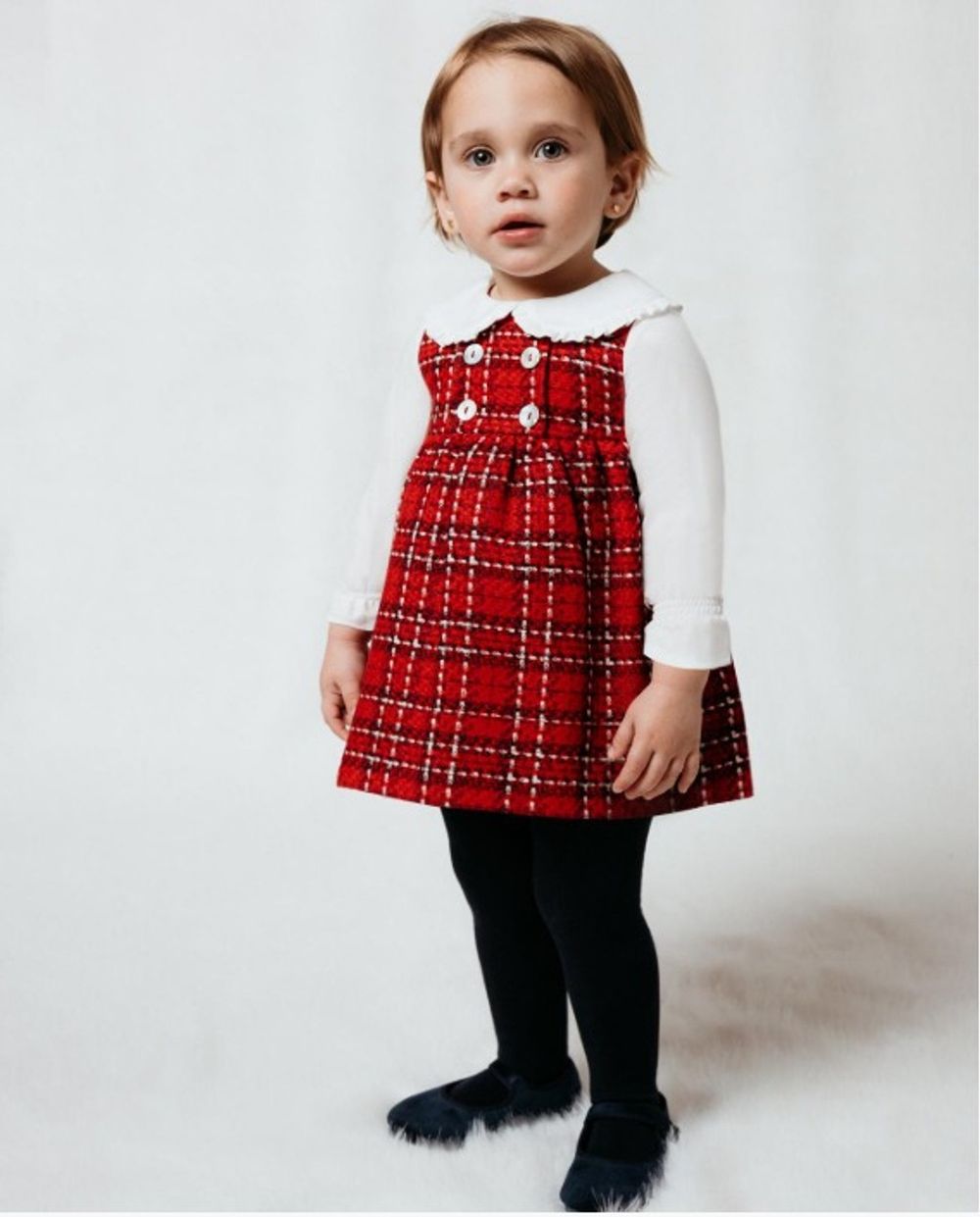 Toddler girl in red plaid pinafore dress over white long-sleeve top with black tights and navy shoes.
