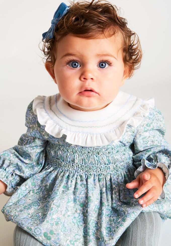 Baby girl in blue floral smocked dress with white ruffle collar and blue bow.