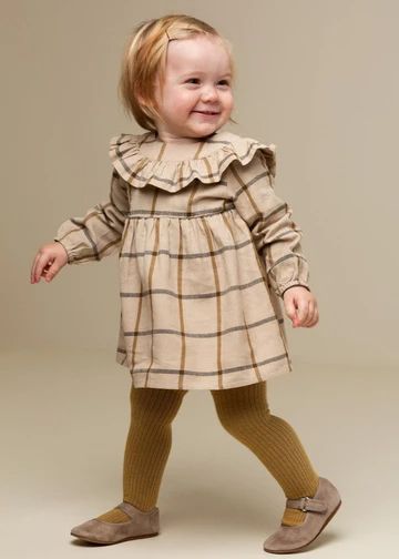 A small child in a beige plaid dress with a ruffled collar, brown tights and mary jane shoes poses adorably.