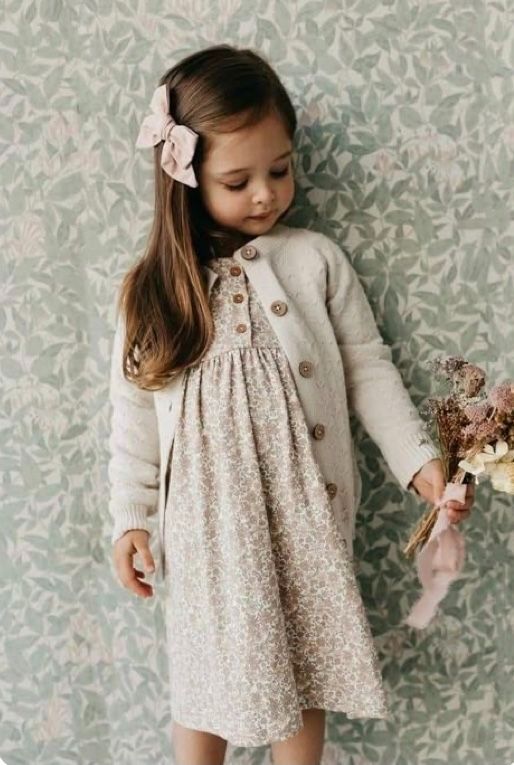 Young child in grey cardigan and floral dress poses against floral wallpaper in vintage style photo series.