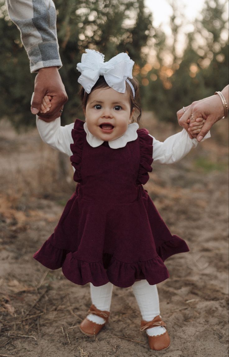 Happy toddler in burgundy dress and white bow holds hands while walking outdoors at sunset.