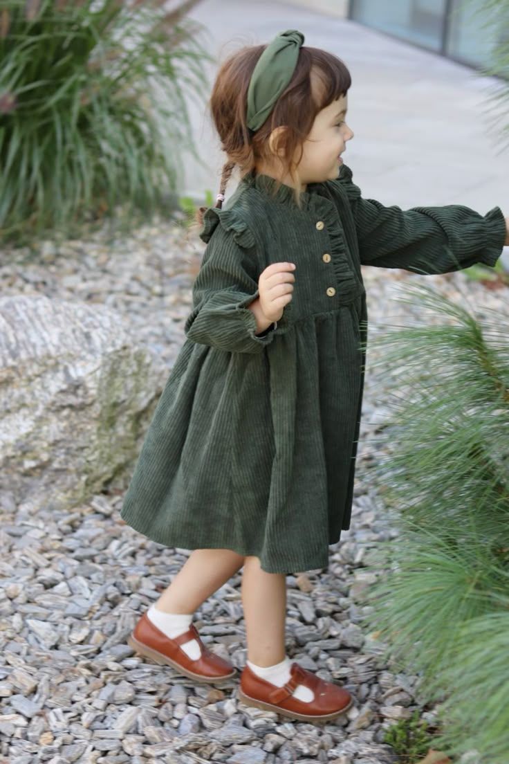 Child wearing green corduroy dress and headband poses playfully among garden plants.