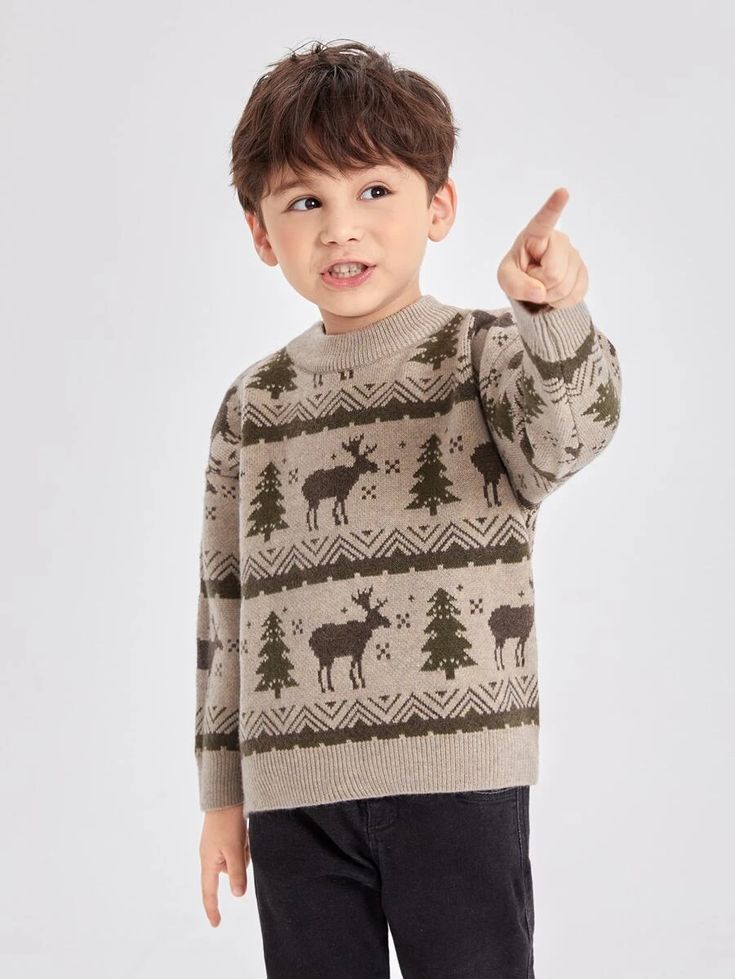 A child in a beige sweater with moose and tree patterns points upward while standing against a light background.